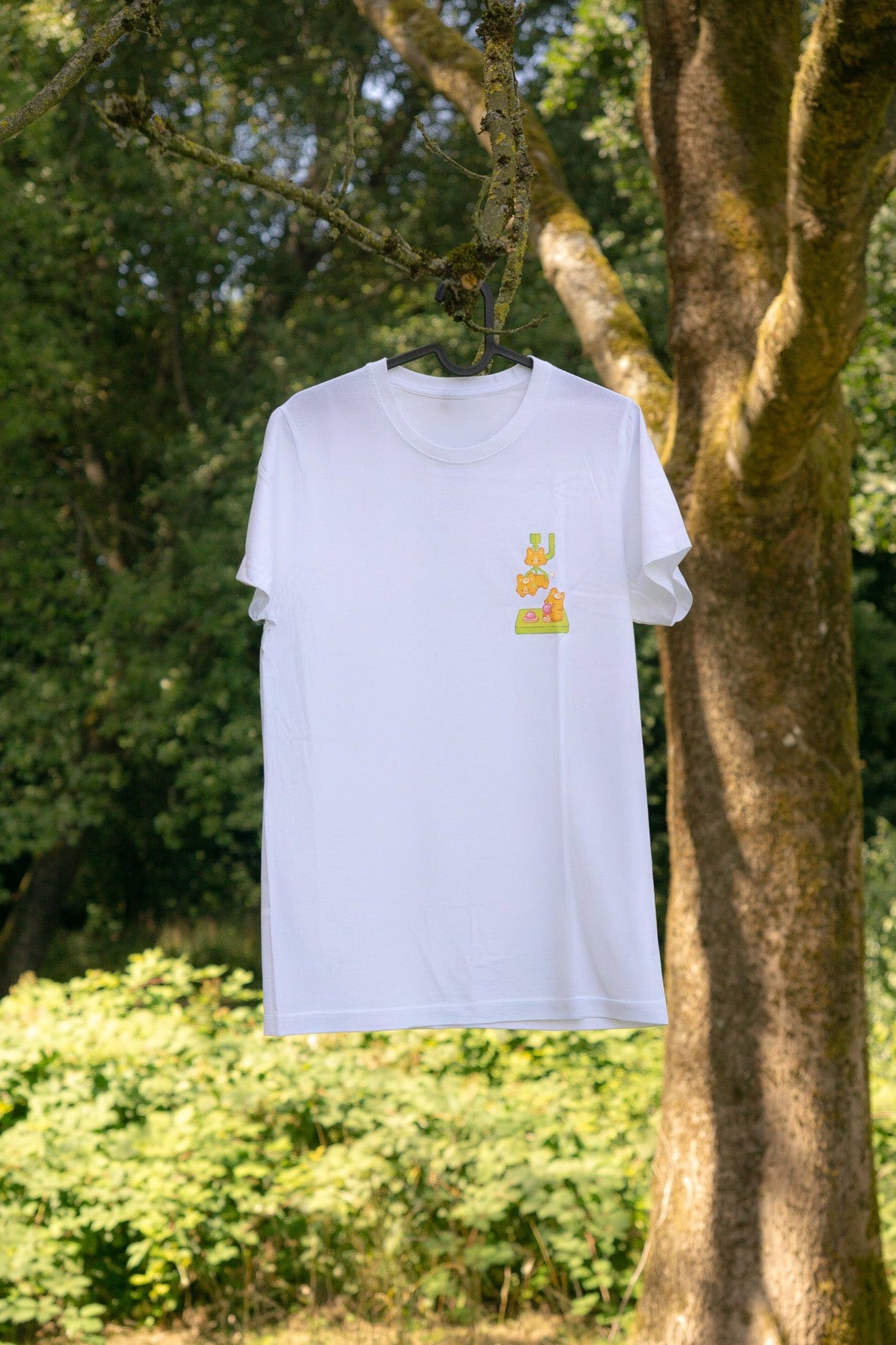 White t-shirt with a small graphic (corgi claw machine) on a hanger against a natural background